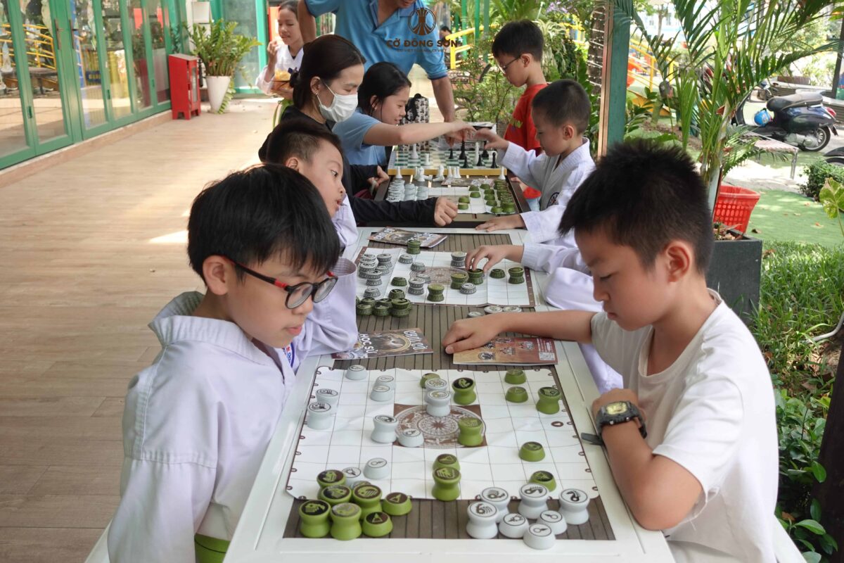 Dãy bàn dài kín chỗ với hình ảnh các bạn nhỏ đang say sưa đấu trí trên bàn Cờ Đông Sơn trước khuôn viên Nhà Sách Thái Hà, Đường Sách TP. Thủ Đức.