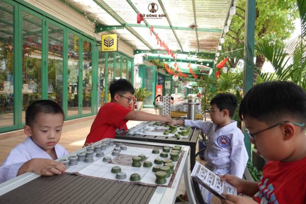 Bốn kỳ thủ nhí, trong đó có hai bé mặc võ phục Karate, đang tập trung cao độ chơi board game Cờ Đông Sơn tại khu vực sinh hoạt trước Nhà Sách Thái Hà, Đường Sách TP. Thủ Đức.
