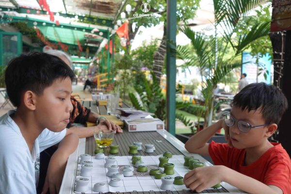 Hình ảnh hai bé trai đang tập trung suy nghĩ nước đi trên bàn Cờ Đông Sơn, bên cạnh là người lớn tuổi đang theo dõi tại Đường sách TP. Thủ Đức