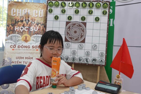 Cận cảnh bé gái đang tập trung cao độ suy nghĩ nước đi trên bàn Cờ Đông Sơn, tay ôm chai nước giải nhiệt tại CLB Lam Sơn.