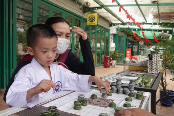 Cận cảnh phụ huynh đang ân cần đồng hành cùng bé trai mặc võ phục tập trung suy nghĩ nước đi Cờ Đông Sơn tại khuôn viên Nhà Sách Thái Hà, Đường Sách TP. Thủ Đức.