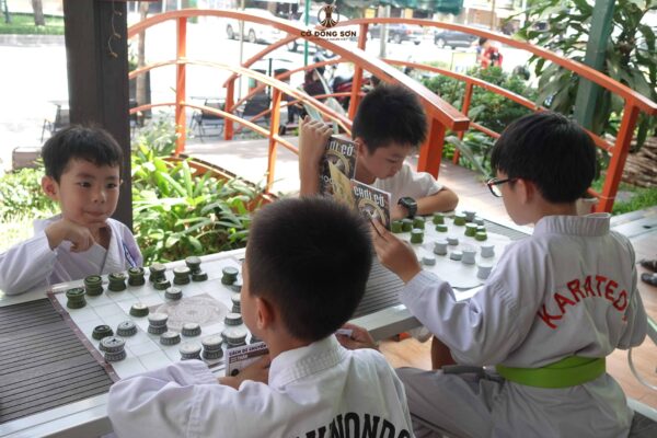 Bốn bé trai mặc trang phục võ thuật đang chăm chú đọc sách hướng dẫn và suy nghĩ nước đi trên bàn Cờ Đông Sơn tại khuôn viên Nhà Sách Thái Hà, Đường Sách TP. Thủ Đức.