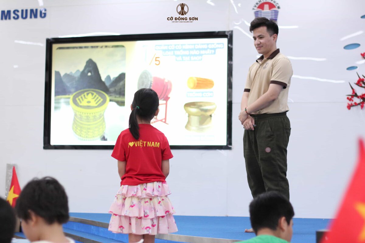 Bé gái mặc áo đỏ "I love Việt Nam" đang tự tin đứng trả lời câu hỏi so sánh hình dáng quân Cờ Đông Sơn với các loại trống truyền thống dưới sự khích lệ của huấn luyện viên tại Khóa hè Cờ Đông Sơn.