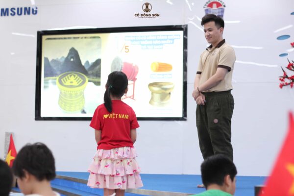 Bé gái mặc áo đỏ "I love Việt Nam" đang tự tin đứng trả lời câu hỏi so sánh hình dáng quân Cờ Đông Sơn với các loại trống truyền thống dưới sự khích lệ của huấn luyện viên tại Khóa hè Cờ Đông Sơn.