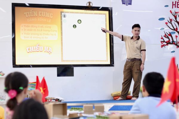 Người hướng dẫn đứng trên sân khấu giới thiệu luật chơi Tiến Cung và Chiếu Hạ trên màn hình LED lớn cho các em học sinh tại Khóa hè Cờ Đông Sơn.