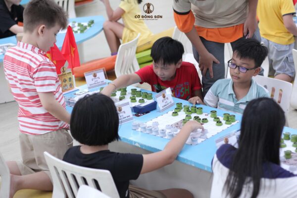 Các kỳ thủ nhí đang quây quần quanh bàn cờ, tập trung cao độ suy nghĩ nước đi trong giờ thực hành thi đấu tại Khóa hè Cờ Đông Sơn dưới sự quan sát của huấn luyện viên.