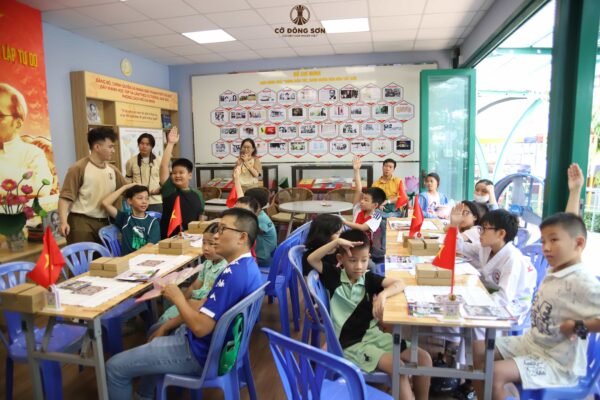 Tập thể các em học sinh hào hứng giơ tay phát biểu trong giờ học tương tác tại Khóa hè Cờ Đông Sơn, với sự quan sát đồng hành của phụ huynh và huấn luyện viên.