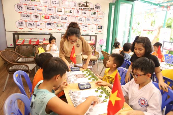 Nữ huấn luyện viên đang tận tình hướng dẫn các kỳ thủ nhí thi đấu trên bàn Cờ Đông Sơn có trang bị đồng hồ tính giờ tại lớp học Khóa hè Cờ Đông Sơn.