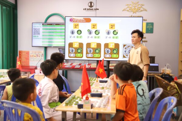 Huấn luyện viên đang đứng trước màn hình lớn hướng dẫn các kỳ thủ nhí cách ghi nhận kết quả và tính điểm ván đấu tại Khóa hè Cờ Đông Sơn.