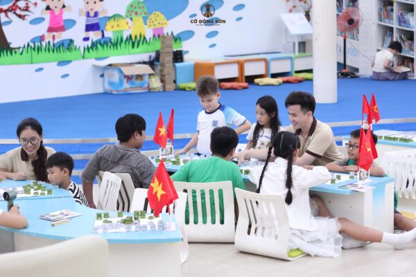 Các huấn luyện viên đang vui vẻ quan sát và hướng dẫn các kỳ thủ nhí quây quần thi đấu giao lưu trên bàn Cờ Đông Sơn tại không gian Khóa hè Cờ Đông Sơn.