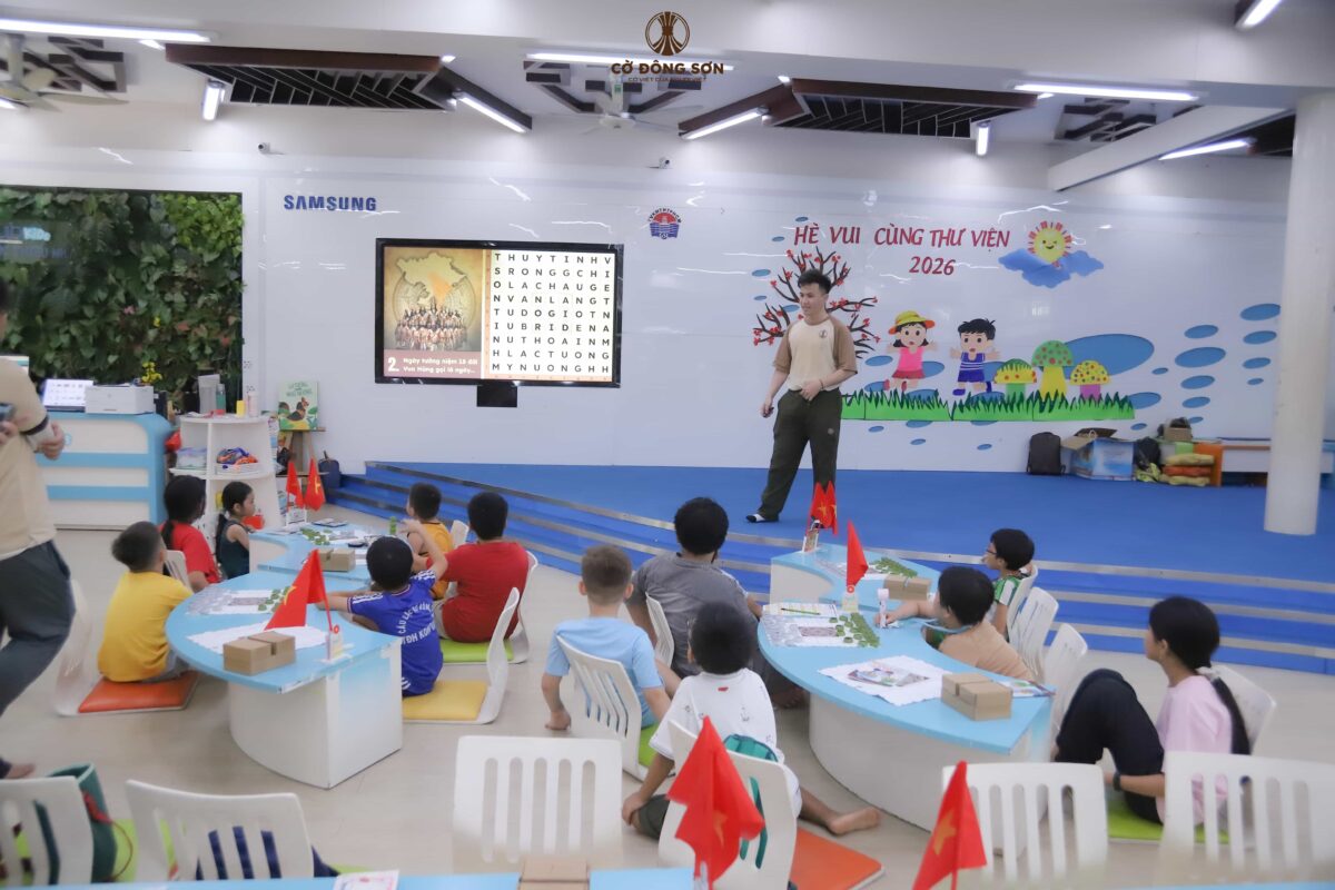 Huấn luyện viên đang hướng dẫn các học viên nhí chơi minigame ô chữ lịch sử trên màn hình lớn tại buổi học thứ hai của Khóa hè Cờ Đông Sơn ở Thư viện.
