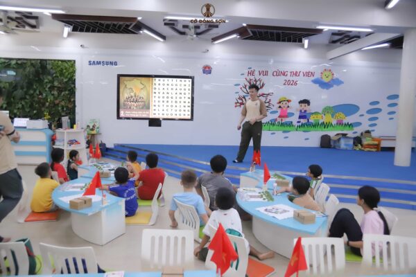Huấn luyện viên đang hướng dẫn các học viên nhí chơi minigame ô chữ lịch sử trên màn hình lớn tại buổi học thứ hai của Khóa hè Cờ Đông Sơn ở Thư viện.