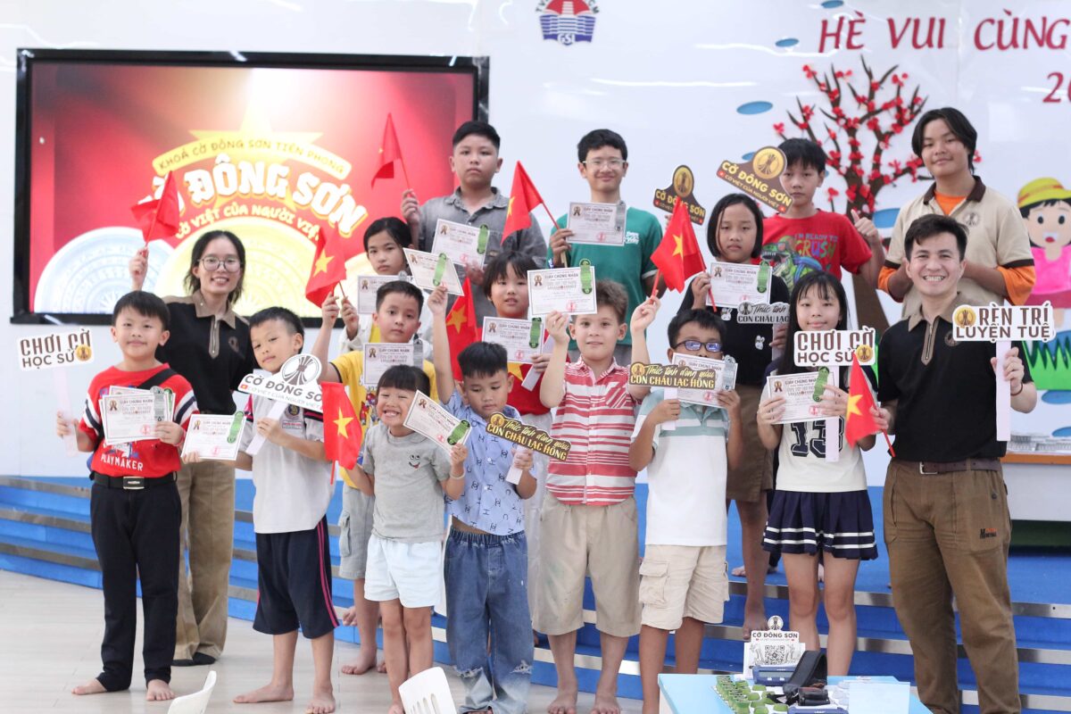 Tập thể kỳ thủ nhí vẫy cờ đỏ sao vàng, tươi cười nhận giấy chứng nhận hoàn thành khóa học từ huấn luyện viên tại Khóa hè Cờ Đông Sơn.