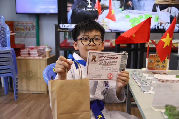 Kỳ thủ nhí rạng rỡ khoe giấy chứng nhận hoàn thành Khóa hè Cờ Đông Sơn 2026 bên bàn Cờ Đông Sơn tại Đường Sách TP. Thủ Đức.