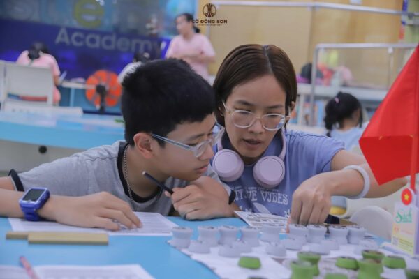 Hình ảnh người mẹ đang chăm chú cùng con trai đọc cẩm nang hướng dẫn và nghiên cứu chiến thuật trên bàn Cờ Đông Sơn trong giờ thực hành của khóa hè.