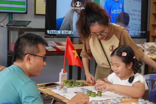 Huấn luyện viên đang mỉm cười tận tình hướng dẫn hai cha con cùng thi đấu trên bàn Cờ Đông Sơn tại không gian Khóa hè Cờ Đông Sơn.