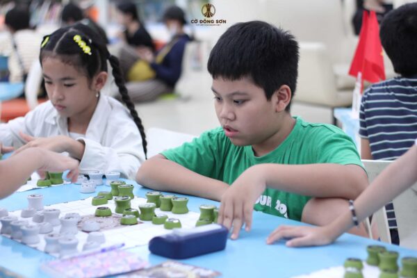 Hình ảnh kỳ thủ nhí mặc áo xanh đang tập trung cao độ suy nghĩ nước đi chiến thuật trên bàn Cờ Đông Sơn trong giờ thi đấu tại Khóa hè Cờ Đông Sơn.