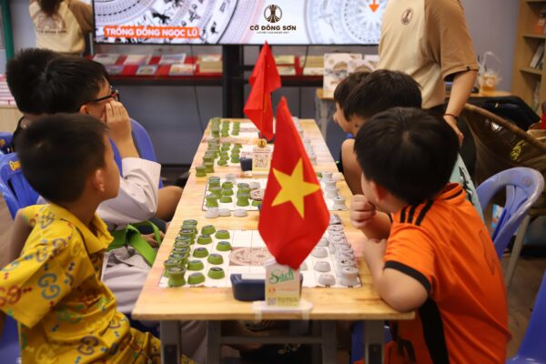 Các kỳ thủ nhí ngồi dọc hai bên bàn Cờ Đông Sơn đang chăm chú theo dõi màn hình bài giảng về họa tiết Trống đồng Ngọc Lũ tại Khóa hè Cờ Đông Sơn.