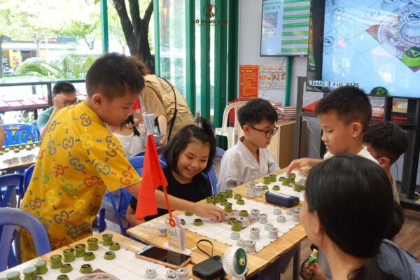 Nhóm các bạn nhỏ đang hào hứng thi đấu thực hành trên bàn Cờ Đông Sơn, nổi bật là cậu bé áo vàng đang đứng lên đi nước cờ chiến thuật trong sự thích thú của bạn bè.