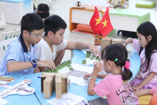 Nhóm các bạn nhỏ nam nữ đang cùng nhau thảo luận, xếp quân và thực hành thi đấu trên bàn Cờ Đông Sơn trong ngày khai mạc khóa hè.